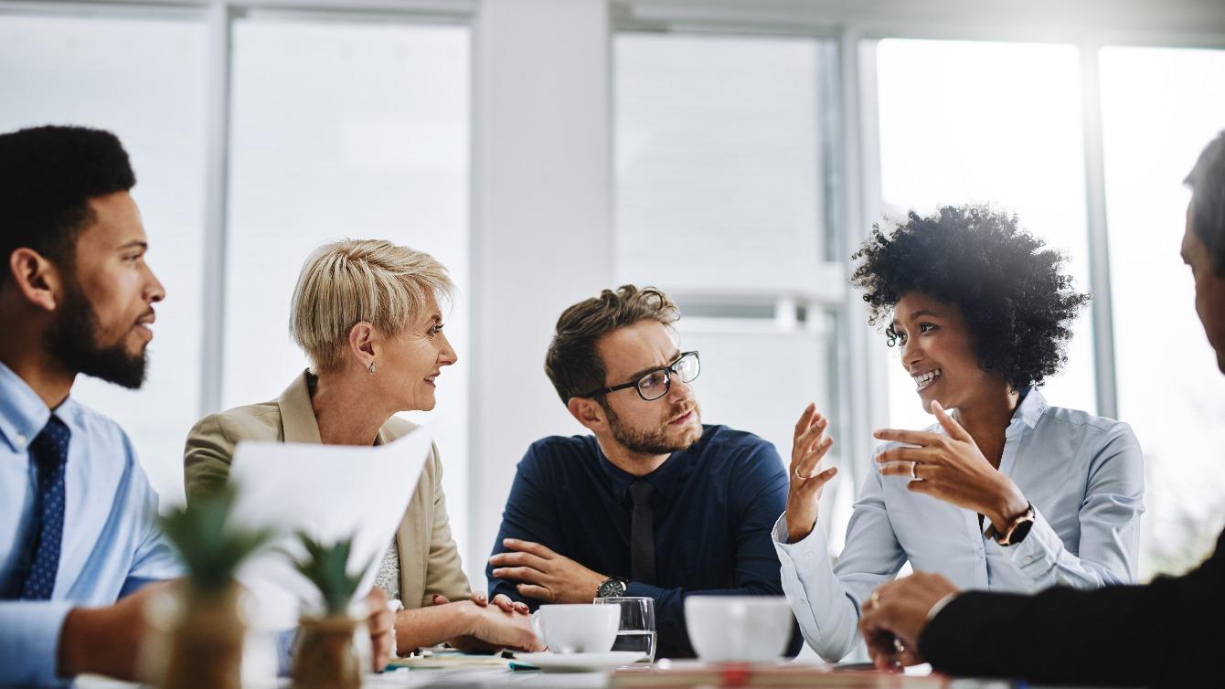 A group of coworkers working together in a meeting room listening to a woman talk over coffee